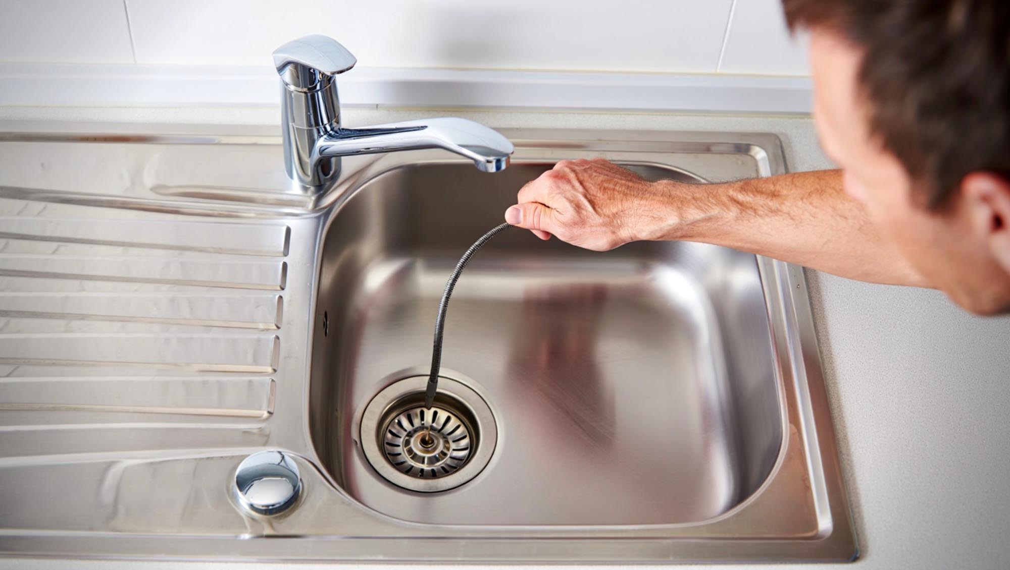 A hand uses a plumbing snake to clear a clogged drain in a white