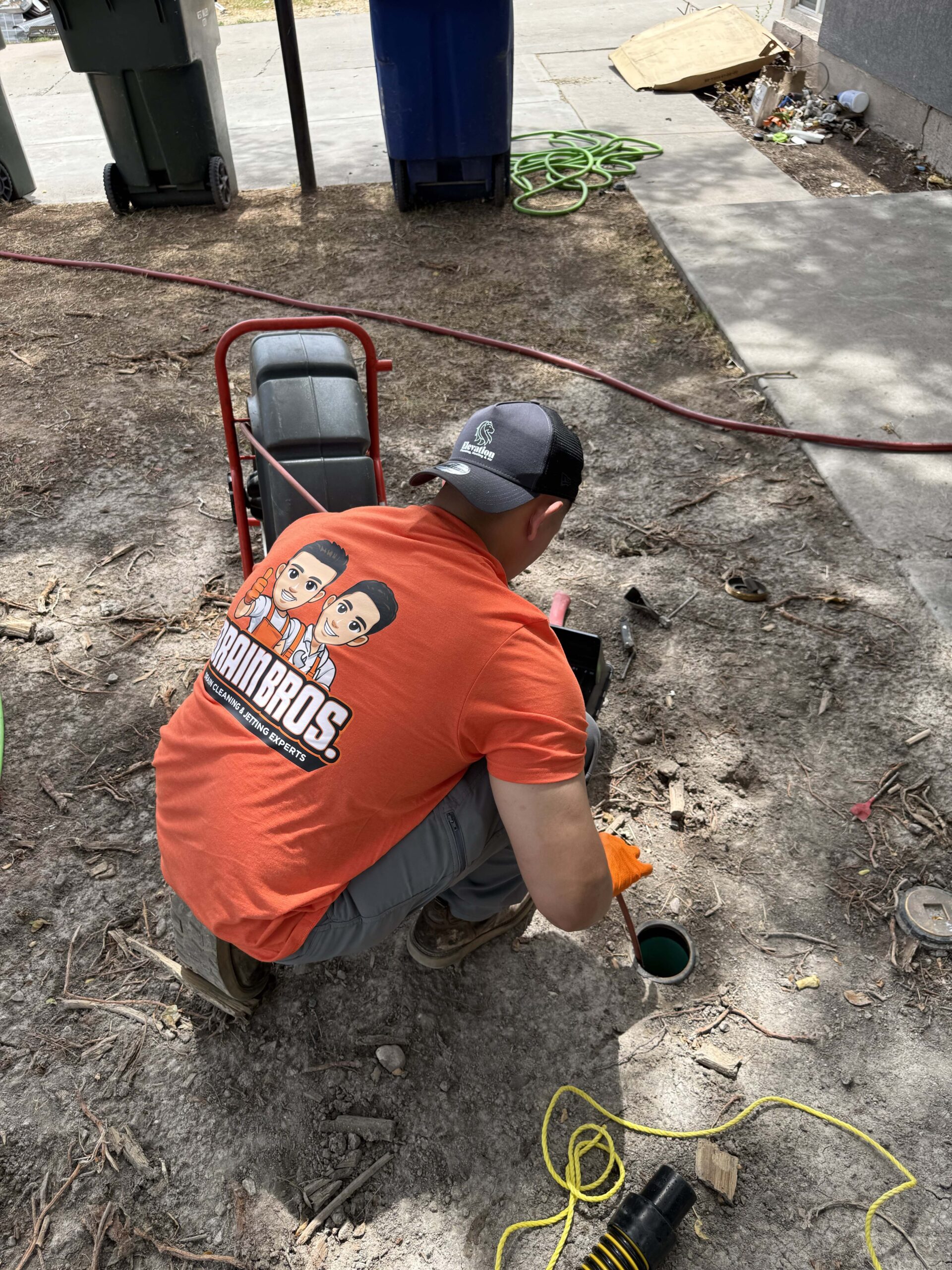 Drain Bros technician crouching down to guide a sewer inspection camera cable into an outdoor drain opening.